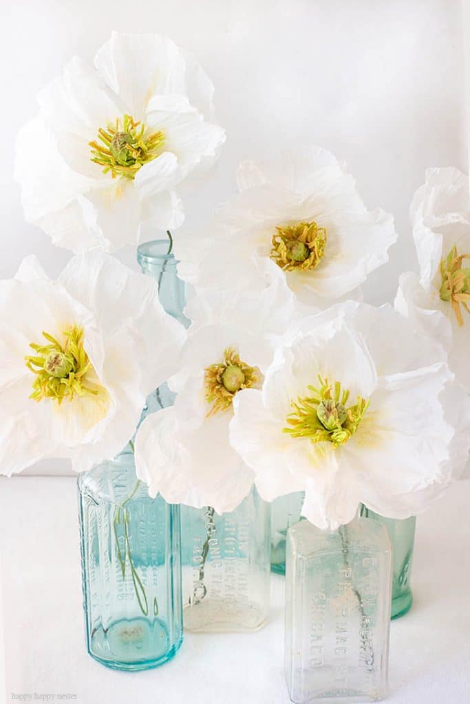 White poppy flowers with yellow centers, my favorite flower inspiration, are arranged in light blue and clear vintage glass bottles against a white background. The image has a soft and delicate feel.