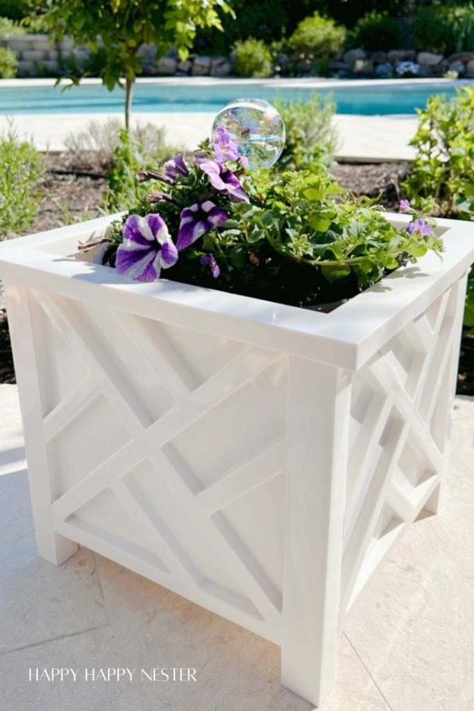 A white geometric planter holds my favorite flower inspiration—purple and white blooms with green foliage. A clear glass globe nestles among the plants. The planter sits on a patio near a pool, with lush greenery and trees in the background.