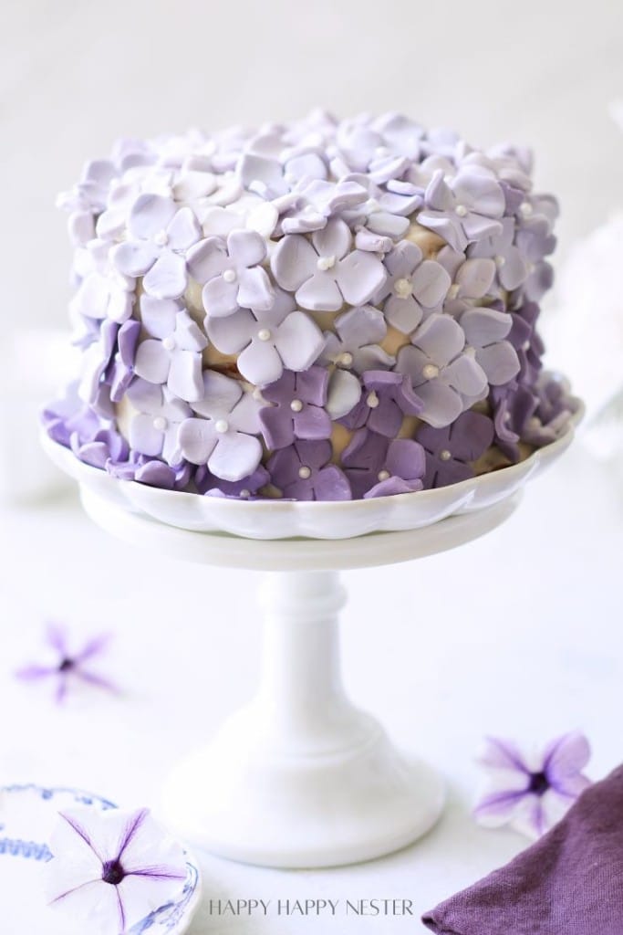 A layered cake decorated with light and dark purple fondant flowers sits on a white cake stand, drawing my favorite flower inspiration, with a few purple flower decorations scattered on the table nearby.