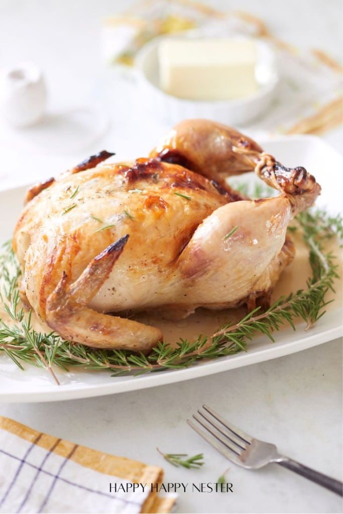 A golden brown, juicy whole roasted chicken garnished with fresh rosemary sits on a white platter—one of my favorite easy dinner ideas. A fork and napkin are nearby, and butter is visible in the background.