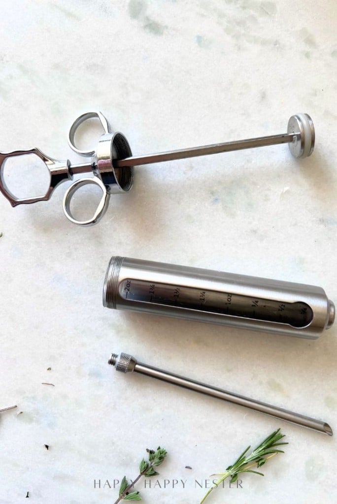 A stainless steel marinade injector disassembled into three parts sits on a white surface next to sprigs of fresh herbs—an essential tool for how to make a tender, juicy steak.