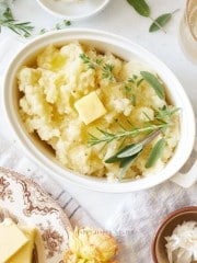 Oval white dish filled with creamy Instant Pot garlic mashed potatoes, topped with a pat of butter and garnished with fresh herbs. Surrounding the dish are plates, butter, a napkin, and a small bowl on a light background.