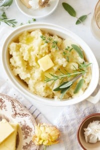 Oval white dish filled with creamy Instant Pot garlic mashed potatoes, topped with a pat of butter and garnished with fresh herbs. Surrounding the dish are plates, butter, a napkin, and a small bowl on a light background.