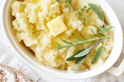 Oval white dish filled with creamy Instant Pot garlic mashed potatoes, topped with a pat of butter and garnished with fresh herbs. Surrounding the dish are plates, butter, a napkin, and a small bowl on a light background.