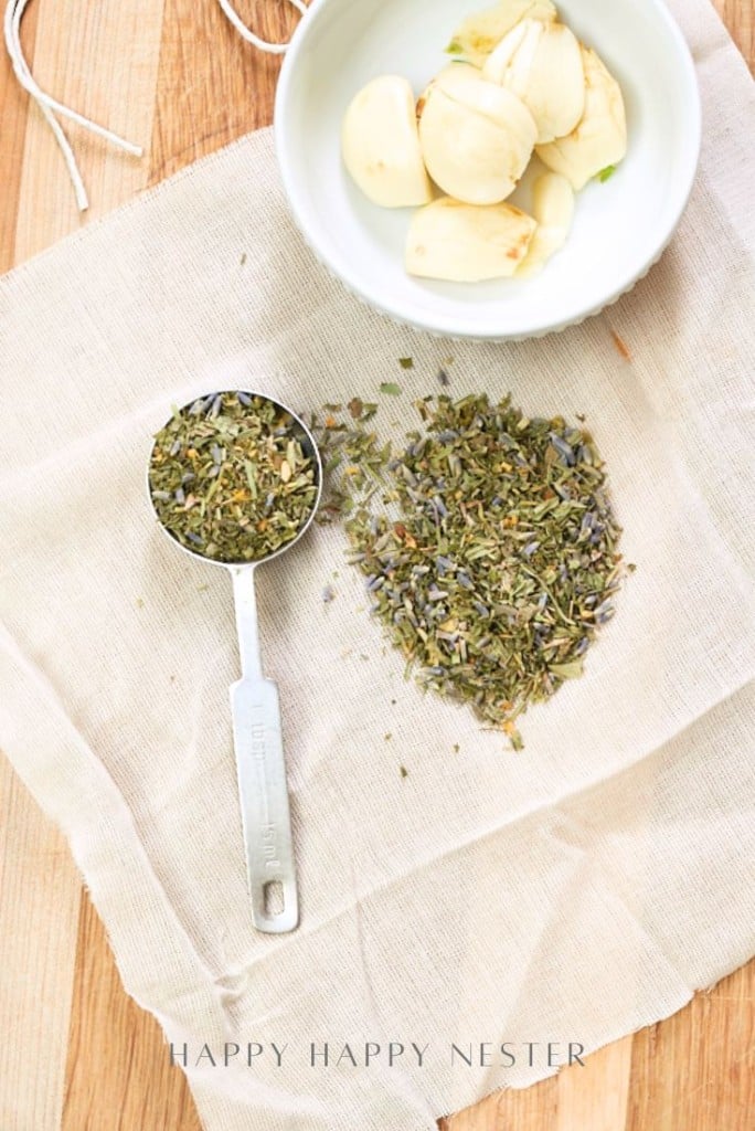 A metal measuring spoon with dried herbs, a small pile of herbs, and a white bowl with peeled garlic cloves—perfect for preparing instant pot garlic mashed potatoes—rest on a beige cloth atop a wooden surface.