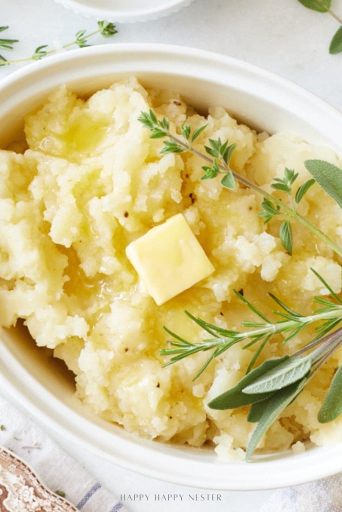 A close-up of creamy Instant Pot garlic mashed potatoes in a white dish, topped with a pat of melting butter and garnished with fresh sprigs of rosemary, sage, and thyme.