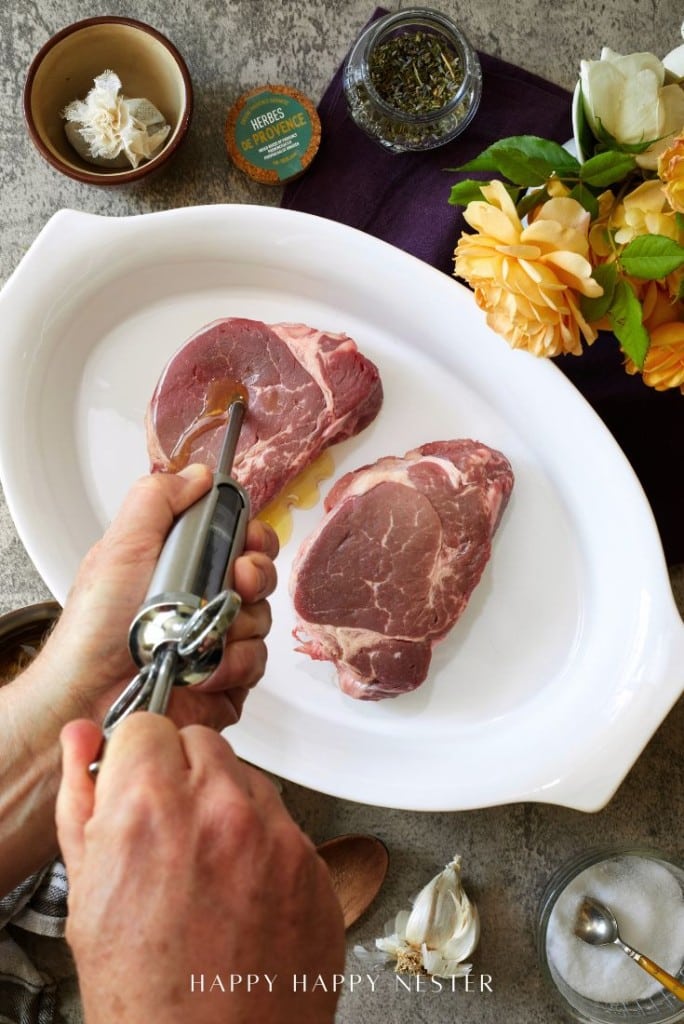 A person demonstrates how to prepare steak by injecting marinade into two raw steaks in a white dish, surrounded by seasonings, garlic, yellow flowers, and kitchen tools on a gray countertop.