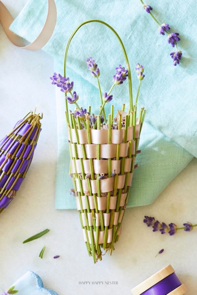 A cone-shaped lavender basket made of woven green and beige ribbon—one of my favorite flower crafts—rests on a light blue cloth, with sprigs of lavender inside and scattered around. Another small woven cone and loose lavender are nearby.