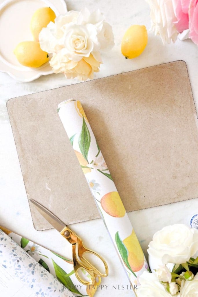 A flat lay of a workspace with easy corkboard placemats, a brown mat, gold scissors, a roll of floral and fruit-patterned paper, white and yellow flowers, and lemons on a marble surface.