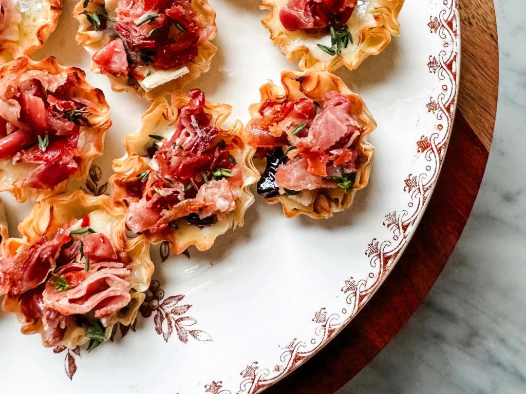 Mini phyllo pastry cups filled with chopped prosciutto, cheese, and herbs are arranged on a white plate with a floral border—perfect for elegant Mother’s Day brunch ideas, set atop a marble surface.