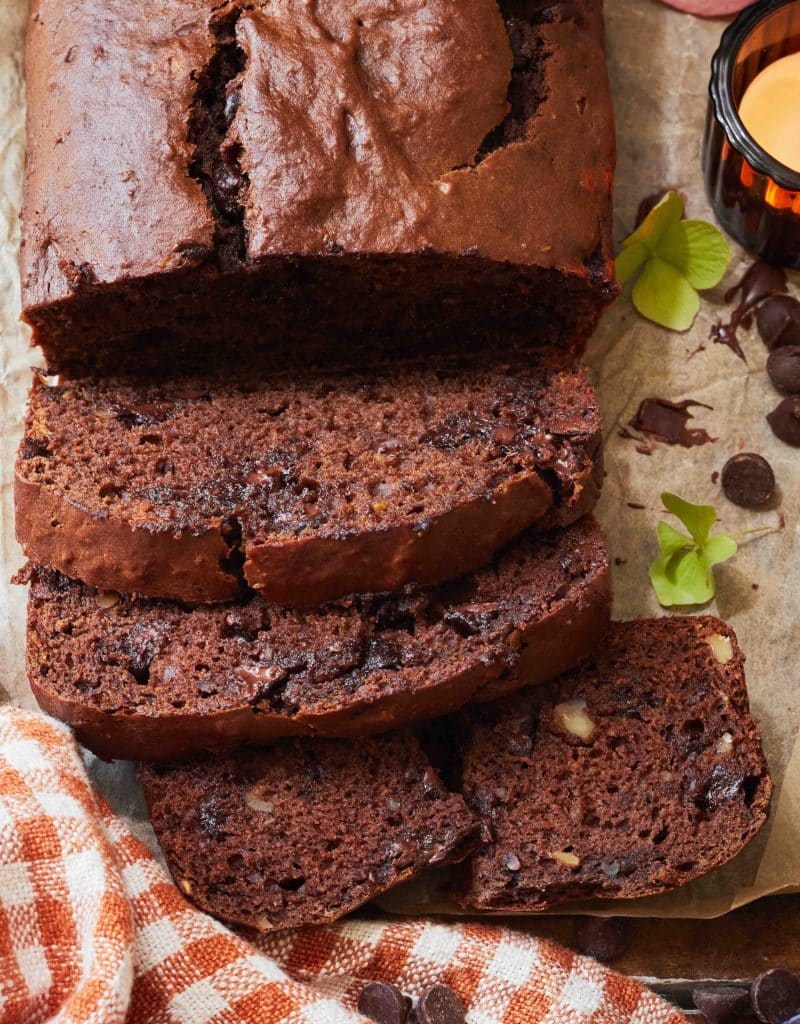 A loaf of chocolate banana bread, one of my favorite chocolate recipes, is sliced on parchment paper, revealing chocolate chips and nuts inside. Nearby are a checkered cloth, a candle, and green leaves for decoration.