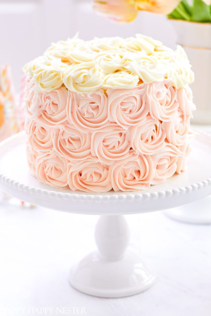A round cake on a white stand, decorated with intricate pink and white buttercream rosettes inspired by my favorite flower. The entire surface is covered in blooms, set against a softly blurred background with hints of pastel colors.