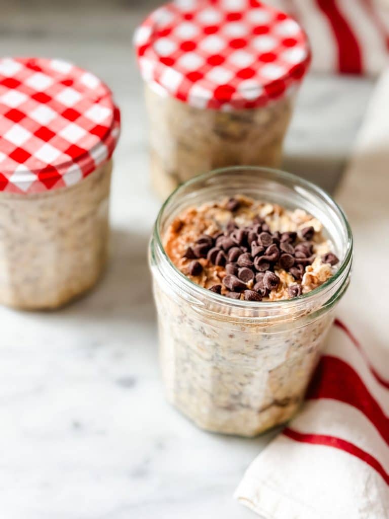 A glass jar filled with overnight oats topped with chocolate chips sits on a marble surface. Two similar jars with red and white checkered lids add charm, perfect for trying your next dessert recipe inspiration.