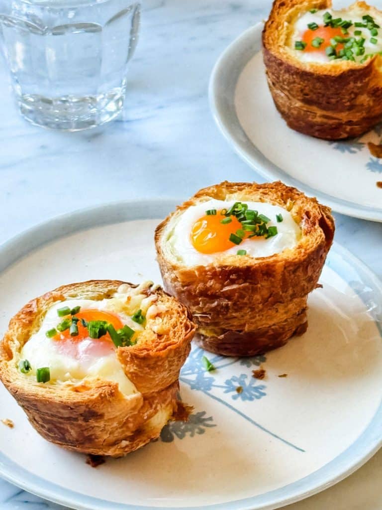 Two croissant muffin cups filled with baked eggs and chopped chives, served on a white plate with a floral design—perfect for mother's day brunch ideas. A glass of water and another plate with more croissant cups are in the background.
