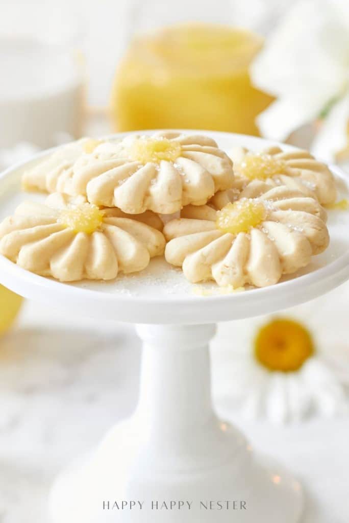 A white cake stand holds several flower-shaped cookies topped with lemon curd and powdered sugar; try this delightful flower cookies recipe. A blurred yellow jar and white flowers are in the background.