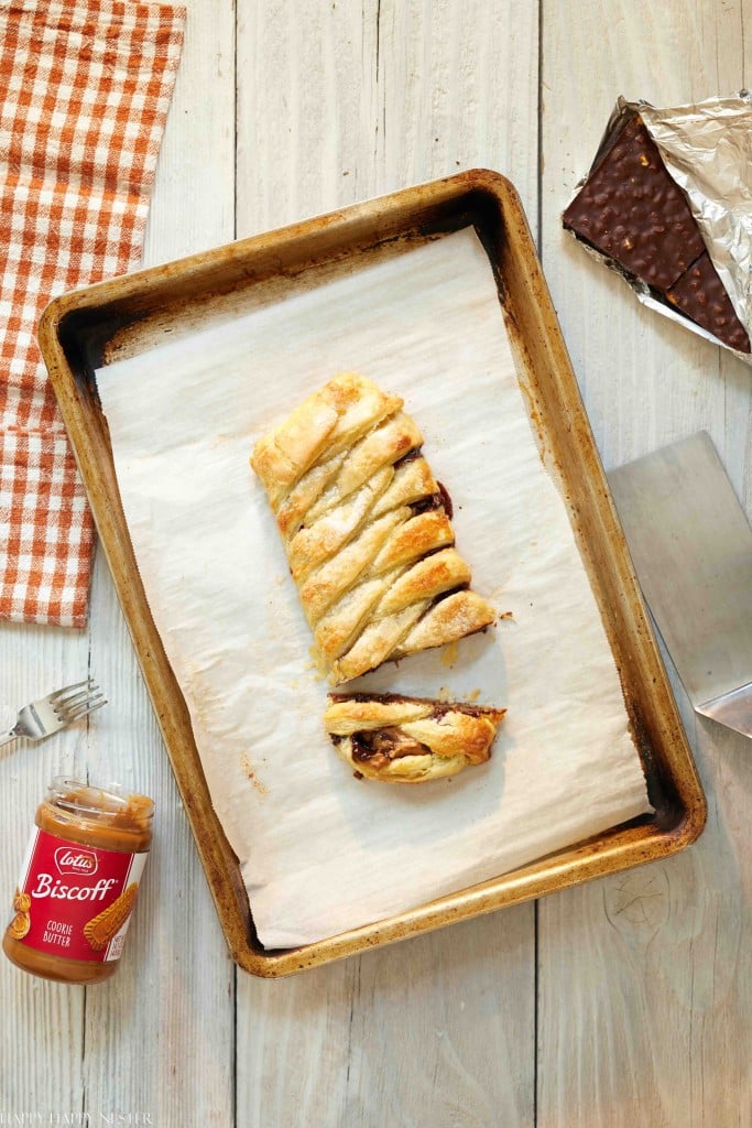 A baked Biscoff pastry loaf on a parchment-lined baking sheet, with one slice cut. Nearby are an open jar of Biscoff spread, a fork, a metal spatula, chocolate—perfect for fans of my favorite chocolate recipes—and an orange checked cloth.
