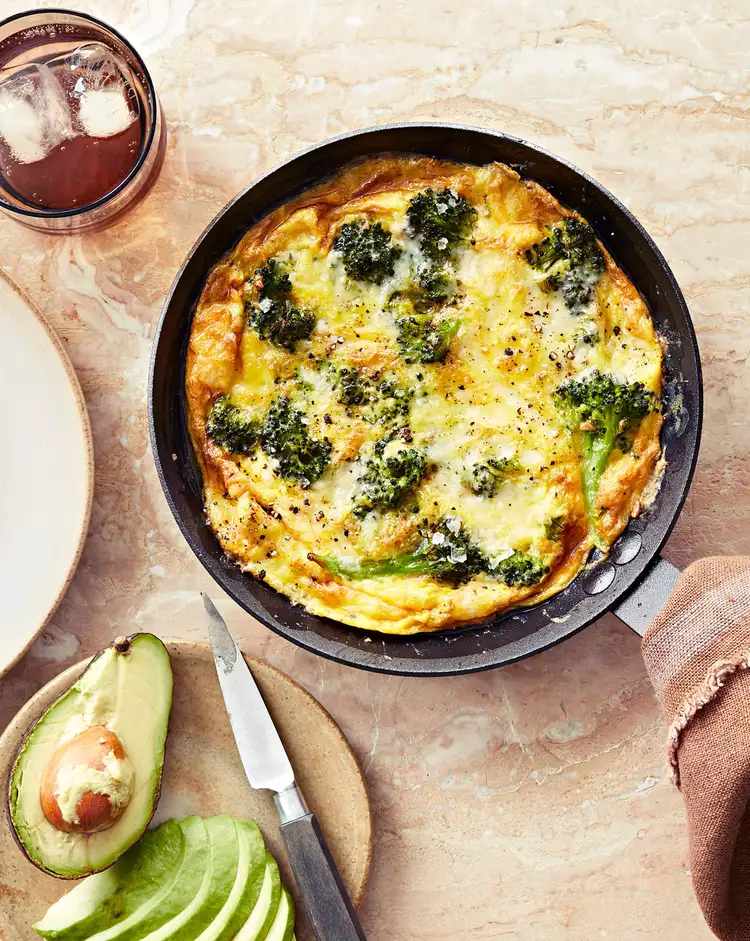 A pan of baked frittata with broccoli sits on a marble surface—perfect for mother’s day brunch ideas. Nearby are a sliced avocado, knife, empty plate, napkin, and a glass of iced drink. The golden frittata is topped with melted cheese.