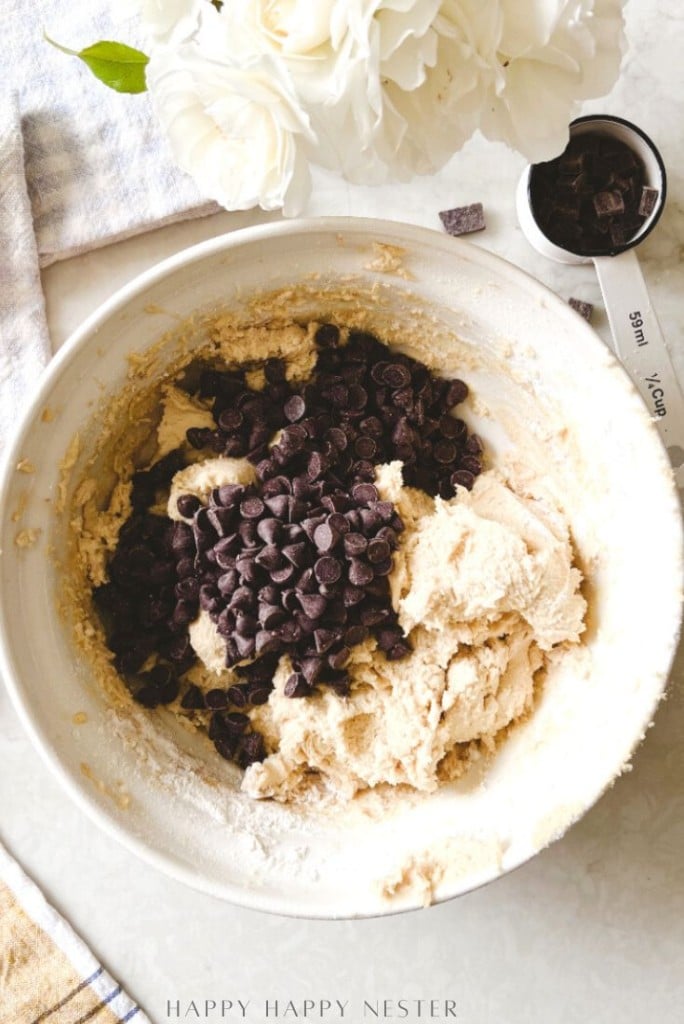 A large white bowl of cookie dough with chocolate chips being mixed in, placed on a light countertop beside a measuring spoon, chocolate chunks, and a vase of white flowers.