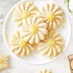 A white plate holds several daisy-shaped sugar cookies with yellow centers, made using a flower cookies recipe, set on a marble surface with more cookies, candles, and floral decor around.