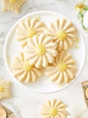 A white plate holds several daisy-shaped sugar cookies with yellow centers, made using a flower cookies recipe, set on a marble surface with more cookies, candles, and floral decor around.