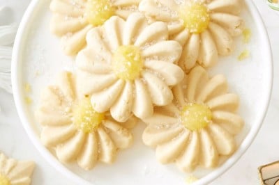 A white plate holds several daisy-shaped sugar cookies with yellow centers, made using a flower cookies recipe, set on a marble surface with more cookies, candles, and floral decor around.