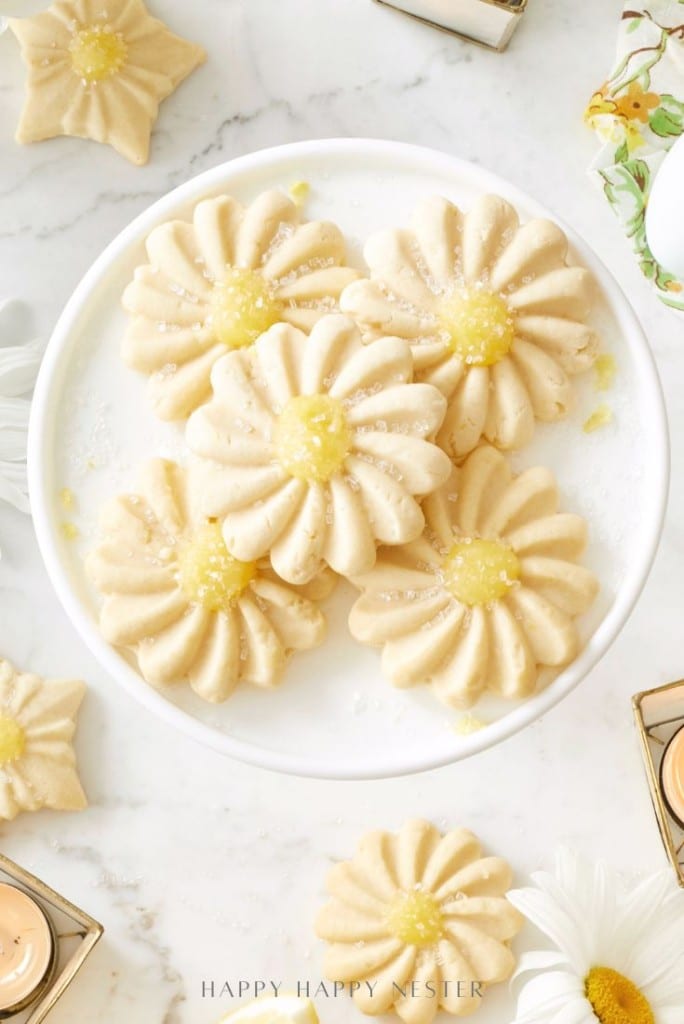 A white plate holds several daisy-shaped sugar cookies with yellow centers, made using a flower cookies recipe, set on a marble surface with more cookies, candles, and floral decor around.
