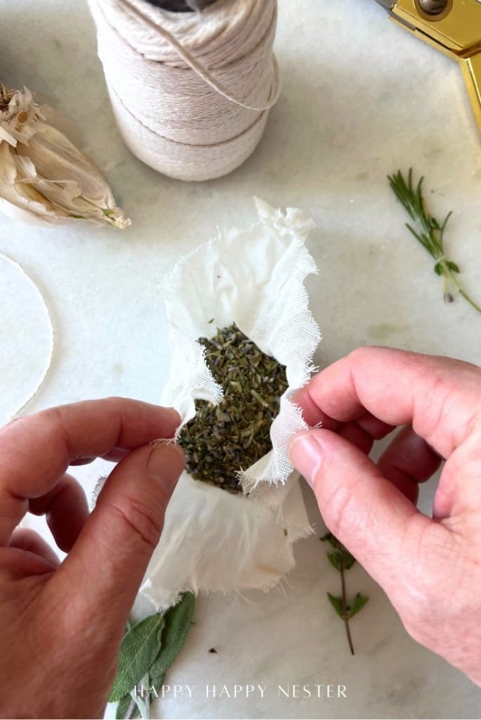 Hands holding a piece of white fabric filled with dried lavender; nearby are twine, scissors, fresh herbs, and garlic—perfect ingredients if you’re also learning how to make a tender juicy steak at home.