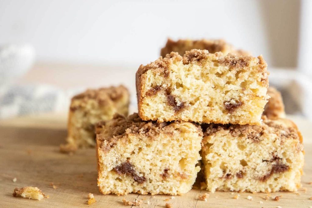 Three pieces of crumb-topped coffee cake with cinnamon swirl are stacked on a wooden surface, making it a delicious addition to your Mother's Day brunch ideas. More slices sit blurred in the background.