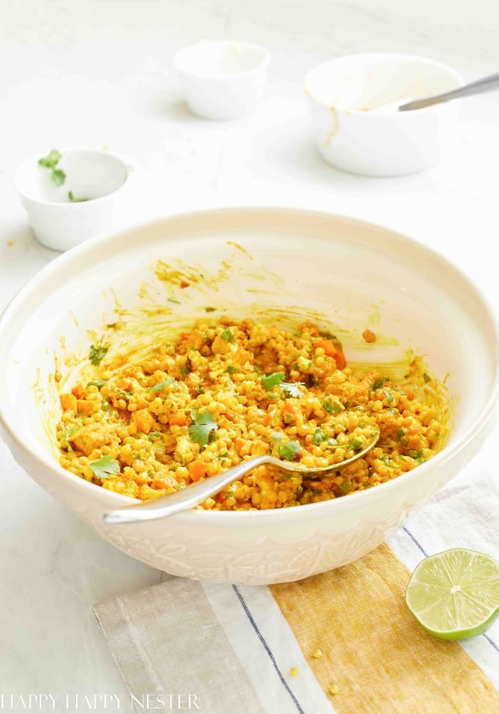 A white mixing bowl filled with a colorful mixture of grains, chopped vegetables, and herbs—one of my favorite side dishes—sits on a table with a spoon inside. A halved lime and small bowls with ingredients are nearby.