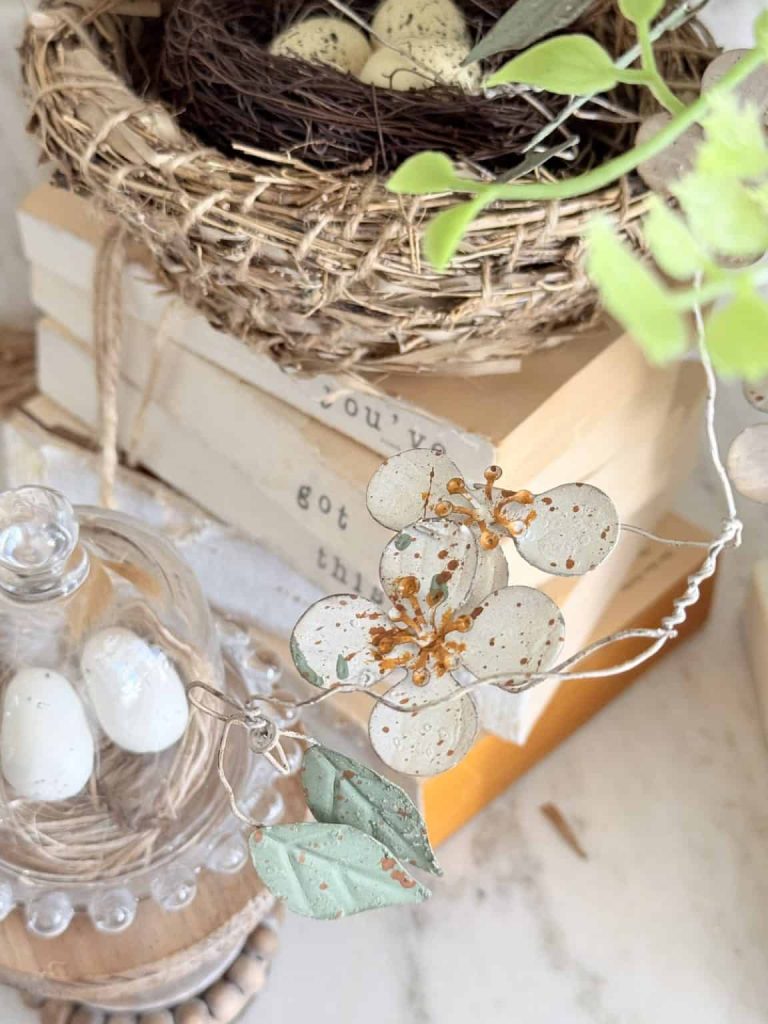 A decorative scene featuring faux bird eggs in a small nest, a stack of books tied with twine, translucent pressed flowers inspired by my favorite flower inspiration, and a glass cloche over more eggs, all set on a marble surface.