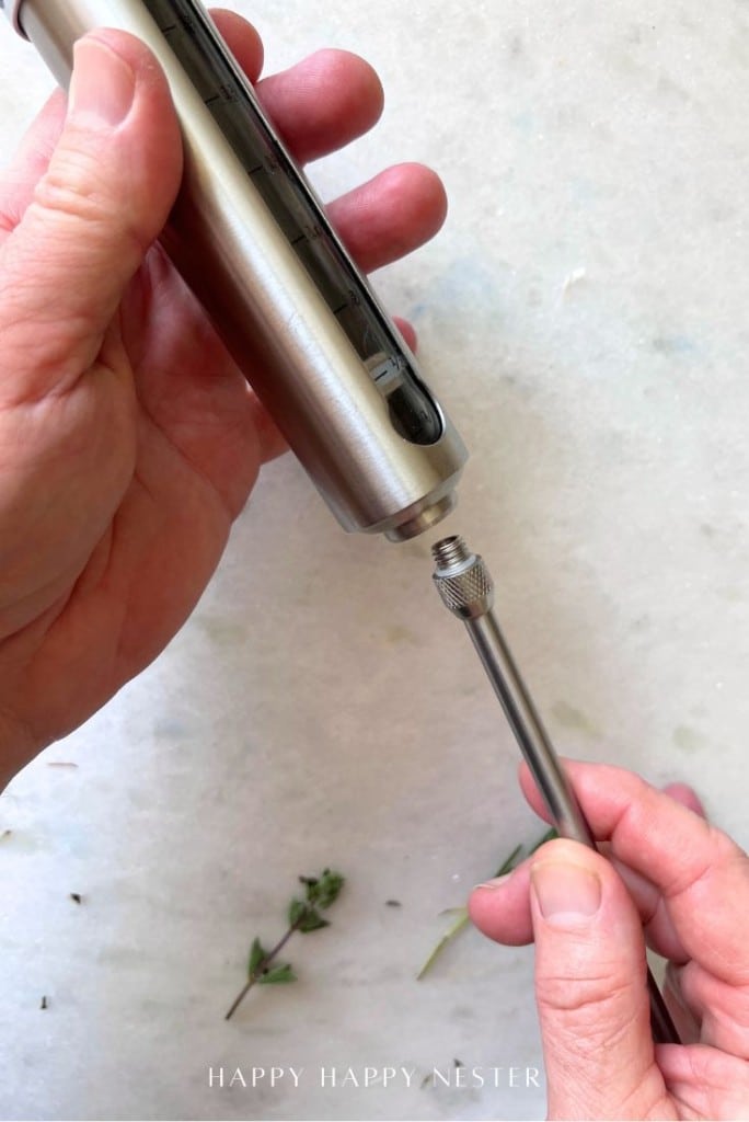 A person attaches a metal needle to a stainless steel marinade injector, preparing herbs below—an essential step in learning how to make a tender, juicy steak.