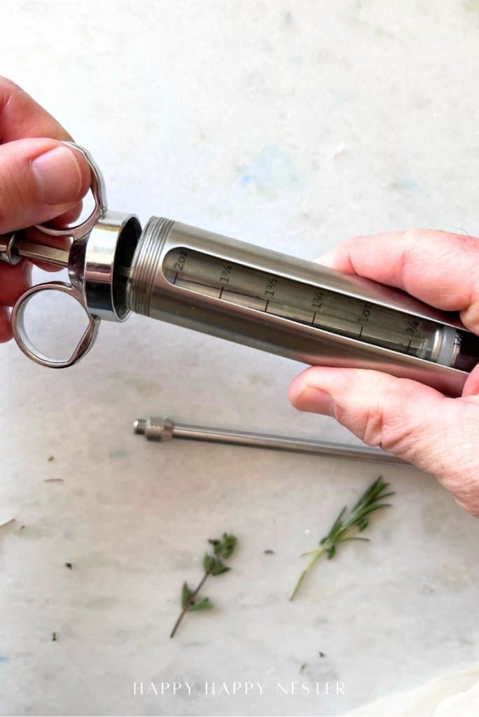 A close-up of hands holding a stainless steel marinade injector with measurement marks—an essential tool for anyone learning how to make a tender juicy steak—next to a detachable needle and fresh herb sprigs on a light surface.