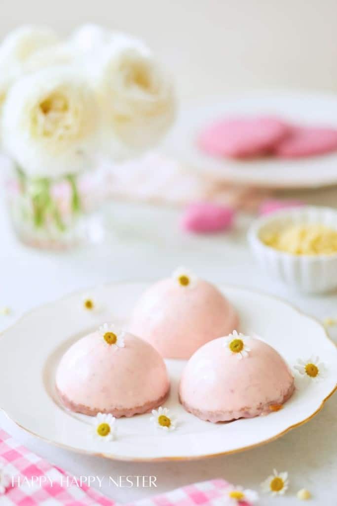 Three dome-shaped pink mousse desserts topped with small white flowers, inspired by my favorite flower inspiration, are displayed on a white plate, with a vase of white roses, pink cookies, and a bowl of white chocolate in the blurred background.