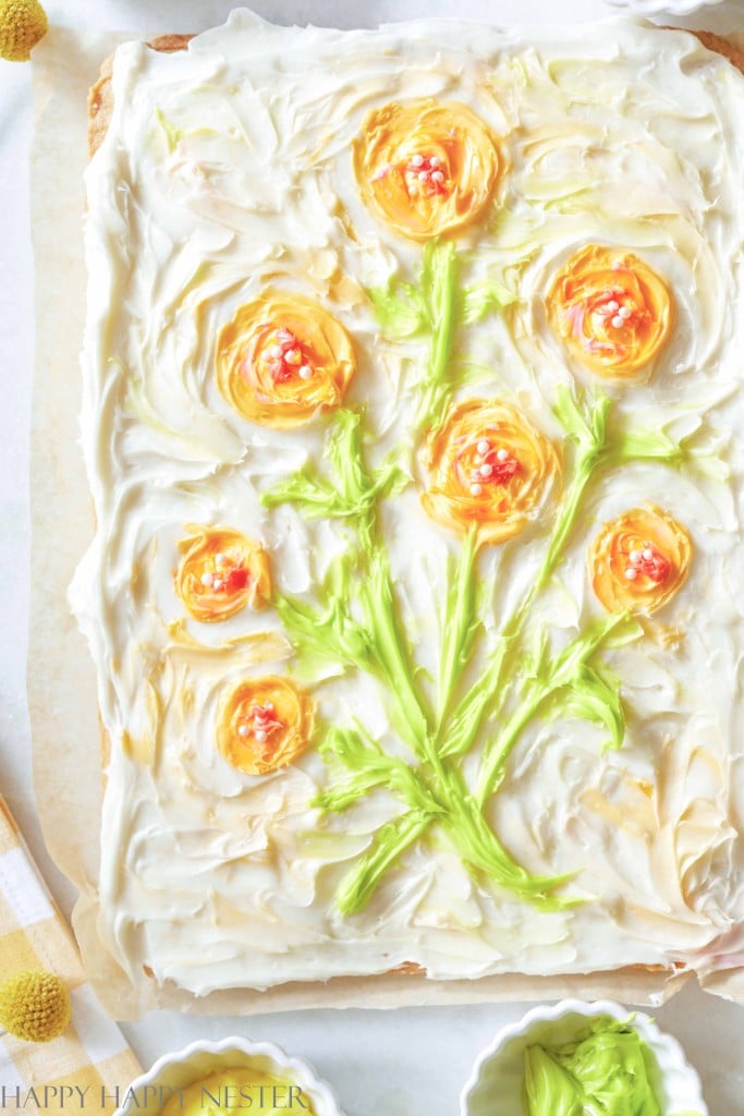 Sheet cake with white frosting decorated to resemble flowers, inspired by my favorite flower inspiration, using yellow and orange icing for the blossoms and green icing for stems and leaves. The cake sits on parchment with bowls and utensils nearby.