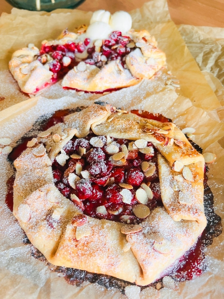 A rustic berry galette topped with sliced almonds and powdered sugar sits on parchment paper—perfect for mother’s day brunch ideas. The crust is golden brown, and the vibrant red berry filling makes it a stunning centerpiece.