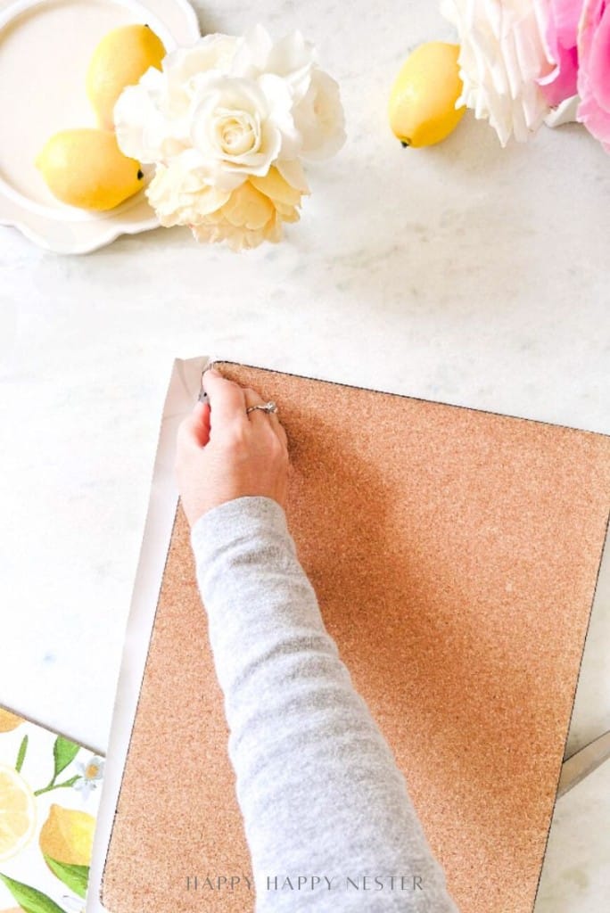 A person’s hand peels paper from easy corkboard placemats on a marble surface, surrounded by lemons and white flowers.