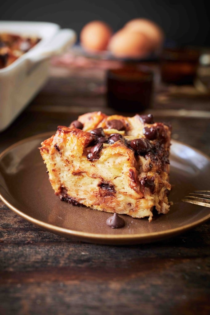 A close-up of a square slice of chocolate chip bread pudding on a brown plate, topped with melted chocolate—one of my favorite chocolate recipes—with eggs and a baking dish blurred in the background.