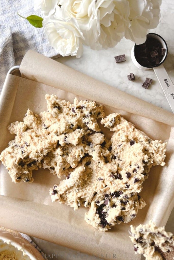 A baking tray lined with parchment paper holds chunky chocolate chip cookie dough, ready to become the perfect chocolate chip bars recipe. Nearby are a measuring cup with chocolate chunks, a metal spoon, and a vase with white flowers on marble.