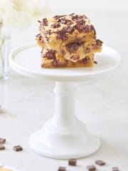 A stack of three chocolate chip bars from a delicious chocolate chip bars recipe is topped with chocolate shavings, sitting on a white cake stand with a blurred floral arrangement in the background.