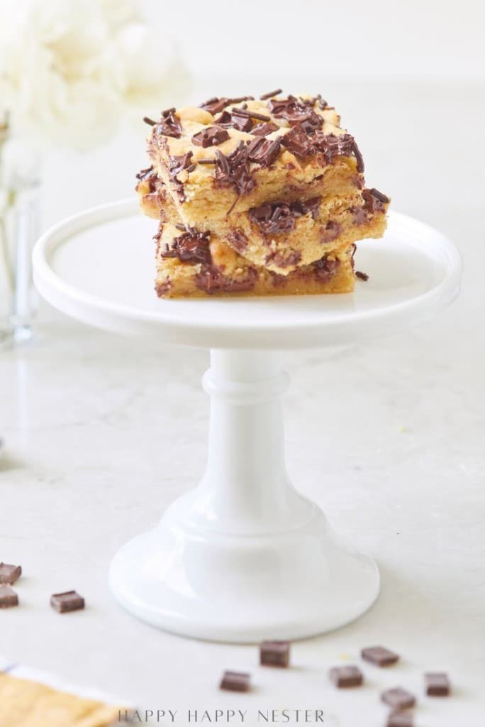 A stack of three chocolate chip bars from a delicious chocolate chip bars recipe is topped with chocolate shavings, sitting on a white cake stand with a blurred floral arrangement in the background.
