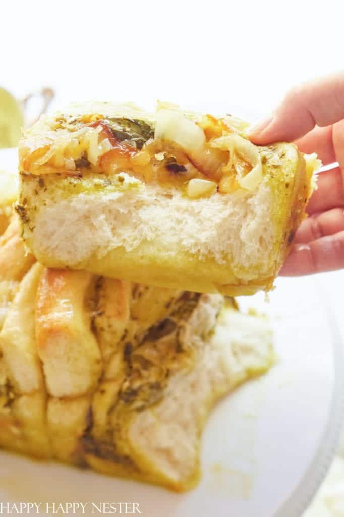 A hand holds a thick, fluffy slice of pull-apart bread filled with cheese, onions, and herbs—one of my favorite side dishes. The bread’s soft texture and golden crust are visible, with more pieces stacked below on a plate.