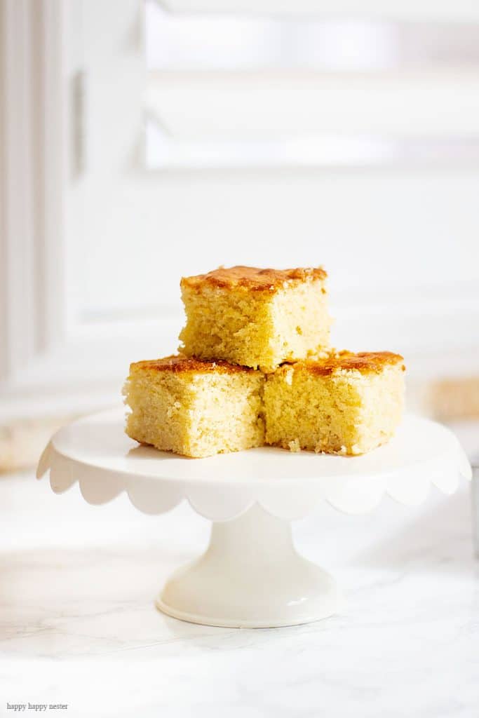 Three square pieces of golden cornbread, one of my favorite side dishes, are stacked on a white cake stand set on a light marble surface in front of a sunlit window.