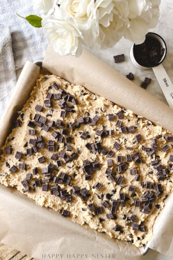 A baking pan lined with parchment paper holds unbaked dough for a chocolate chip bars recipe, topped with chocolate chunks. A white flower bouquet and a metal measuring cup filled with more chocolate chunks are nearby.