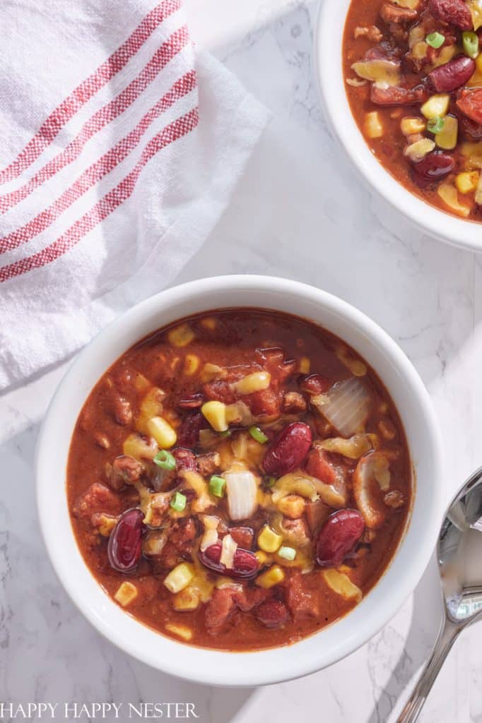 A bowl of chili topped with corn, kidney beans, green onions, and diced onions sits on a marble surface—one of my favorite easy dinner ideas—next to a spoon, with a red-striped kitchen towel and part of another bowl visible.
