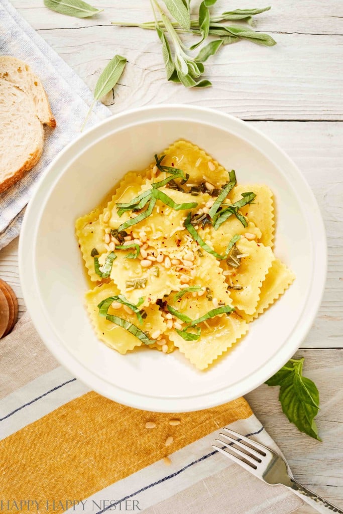A white bowl filled with ravioli, topped with fresh basil, pine nuts, and grated cheese, makes one of my favorite easy dinner ideas. The bowl sits on a wooden table with a slice of bread, fresh herbs, and a yellow-striped napkin beside it.