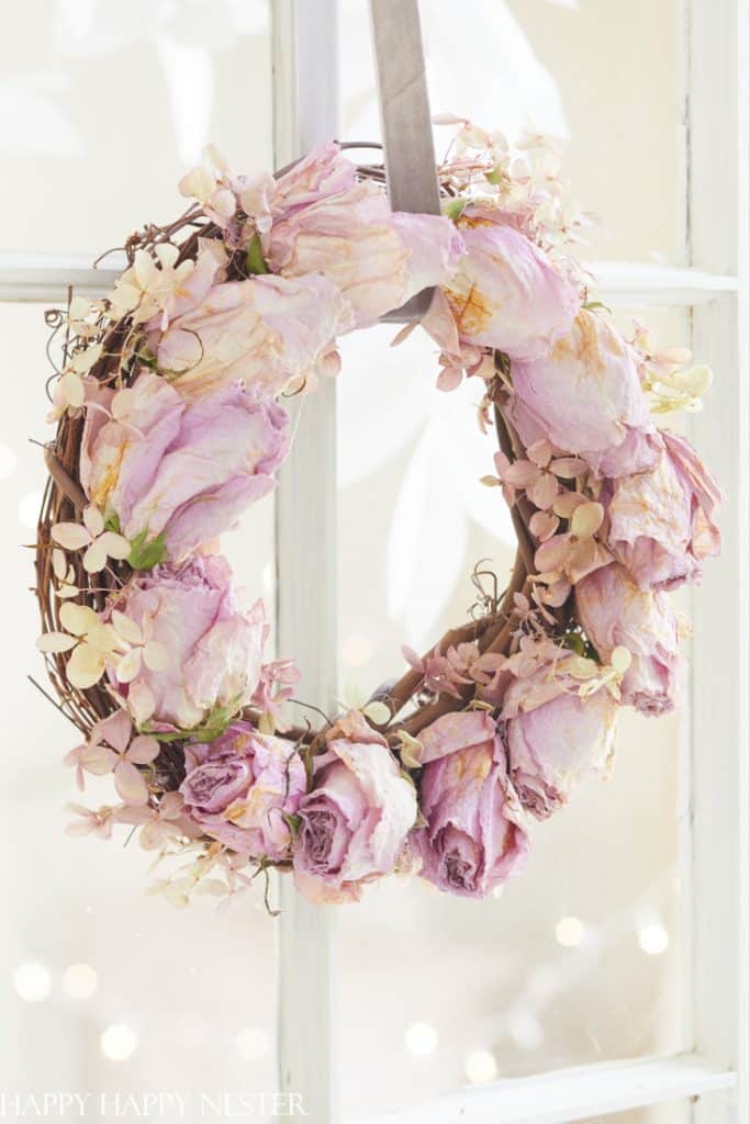 A wreath made of pale pink dried roses and small flowers—one of my favorite flower crafts—hangs by a gray ribbon on a window, with soft natural light streaming through the glass.