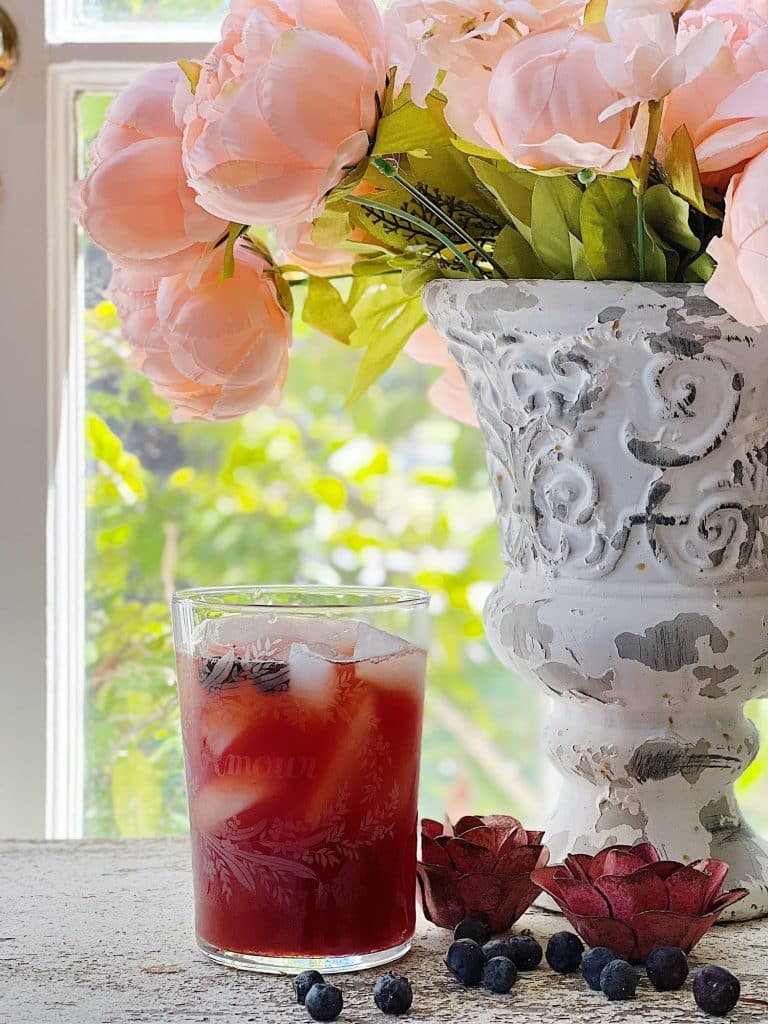 A glass of iced red juice, one of my favorite summer drinks, sits on a rustic white table beside scattered blueberries and two decorative flowers. In the background, sunlight streams through a window, illuminating a white ornate vase of pink peonies.