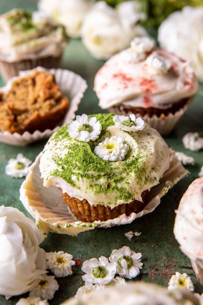 A cupcake topped with white frosting, green matcha powder, and small white flowers sits on an open wrapper—one of my favorite floral desserts. More cupcakes and delicate blooms are blurred in the background.