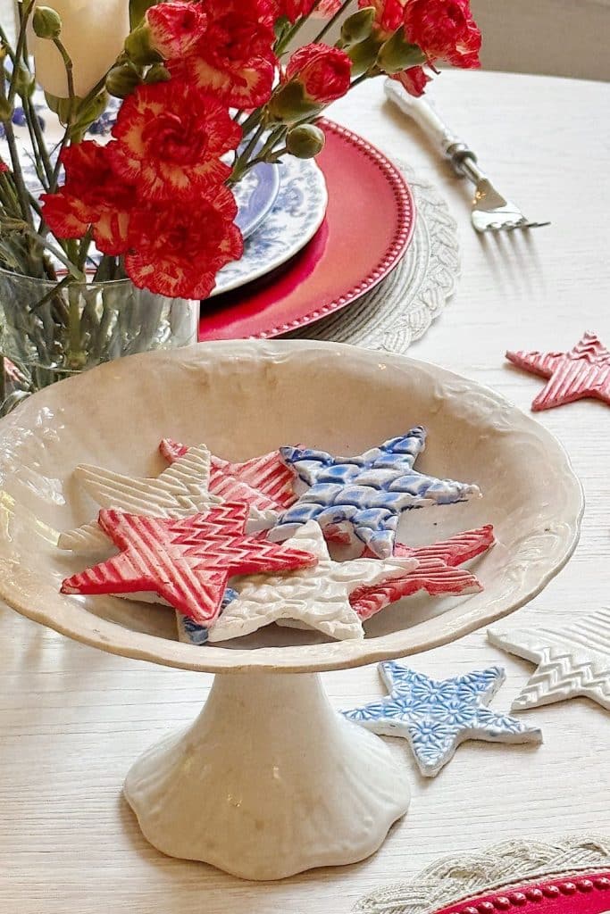 A white pedestal dish holds decorative star-shaped ornaments in red, white, and blue patterns. Red flowers, a red-rimmed plate, and a fork are seen on a white table in the background.