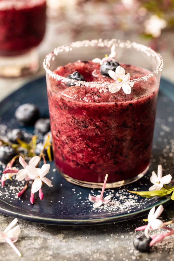 A glass of dark berry slush—one of my favorite summer drinks—garnished with fresh blueberries and white jasmine flowers, with a sugared rim. The glass sits on a dark plate adorned with more berries, jasmine flowers, and sugar.